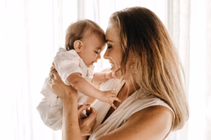Smiling mother holds her laughing baby girl in a bright, sunlit room at home.