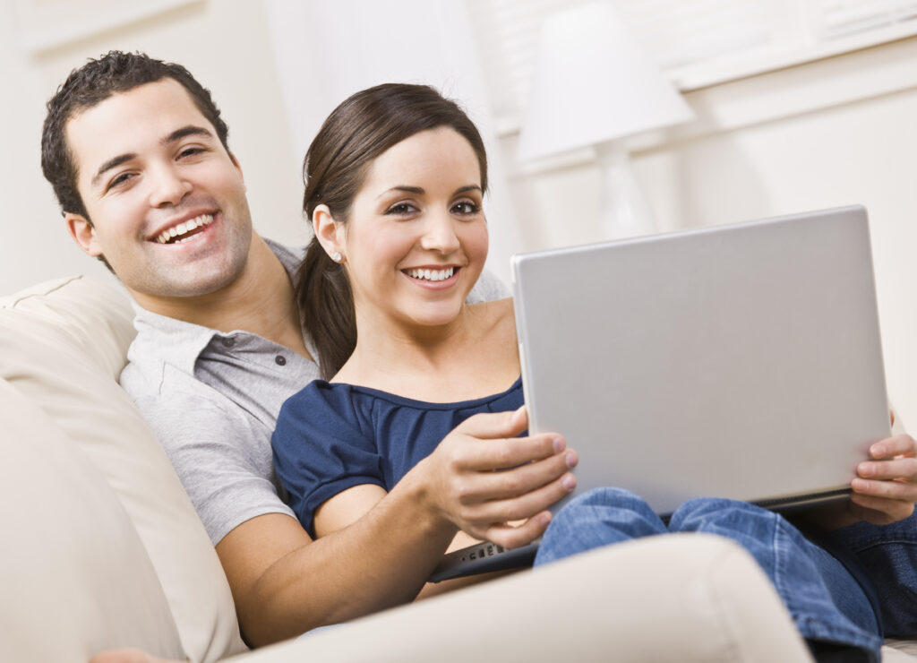 An attractive young couple sitting on a couch together and holding a laptop. They are smiling directly at the camera. Horizontally framed shot.