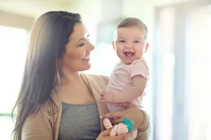 Smiling mother holds her laughing baby girl in a bright, sunlit room at home.