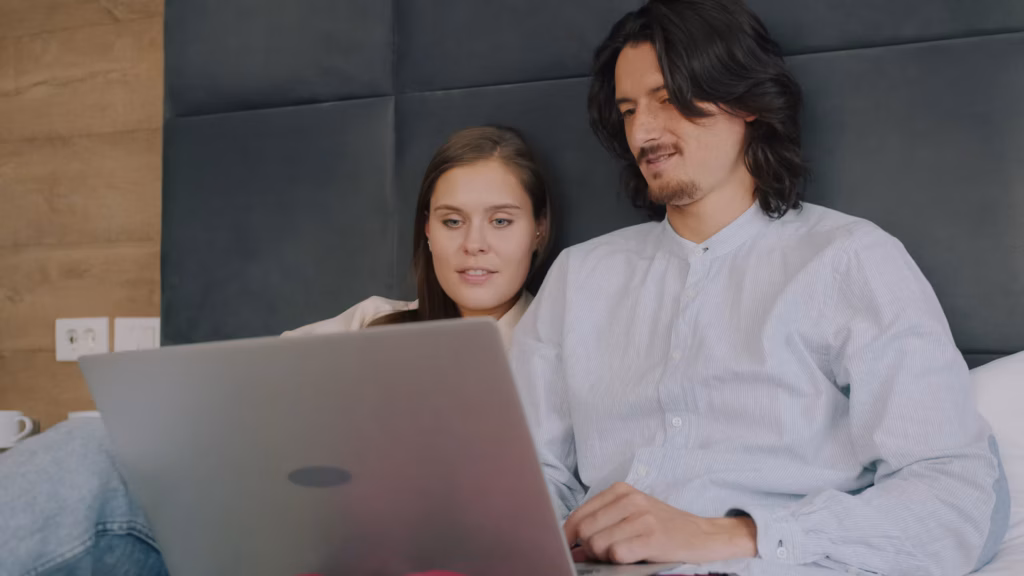 An attractive young couple sitting on a couch together and holding a laptop. They are smiling directly at the camera. Horizontally framed shot.
